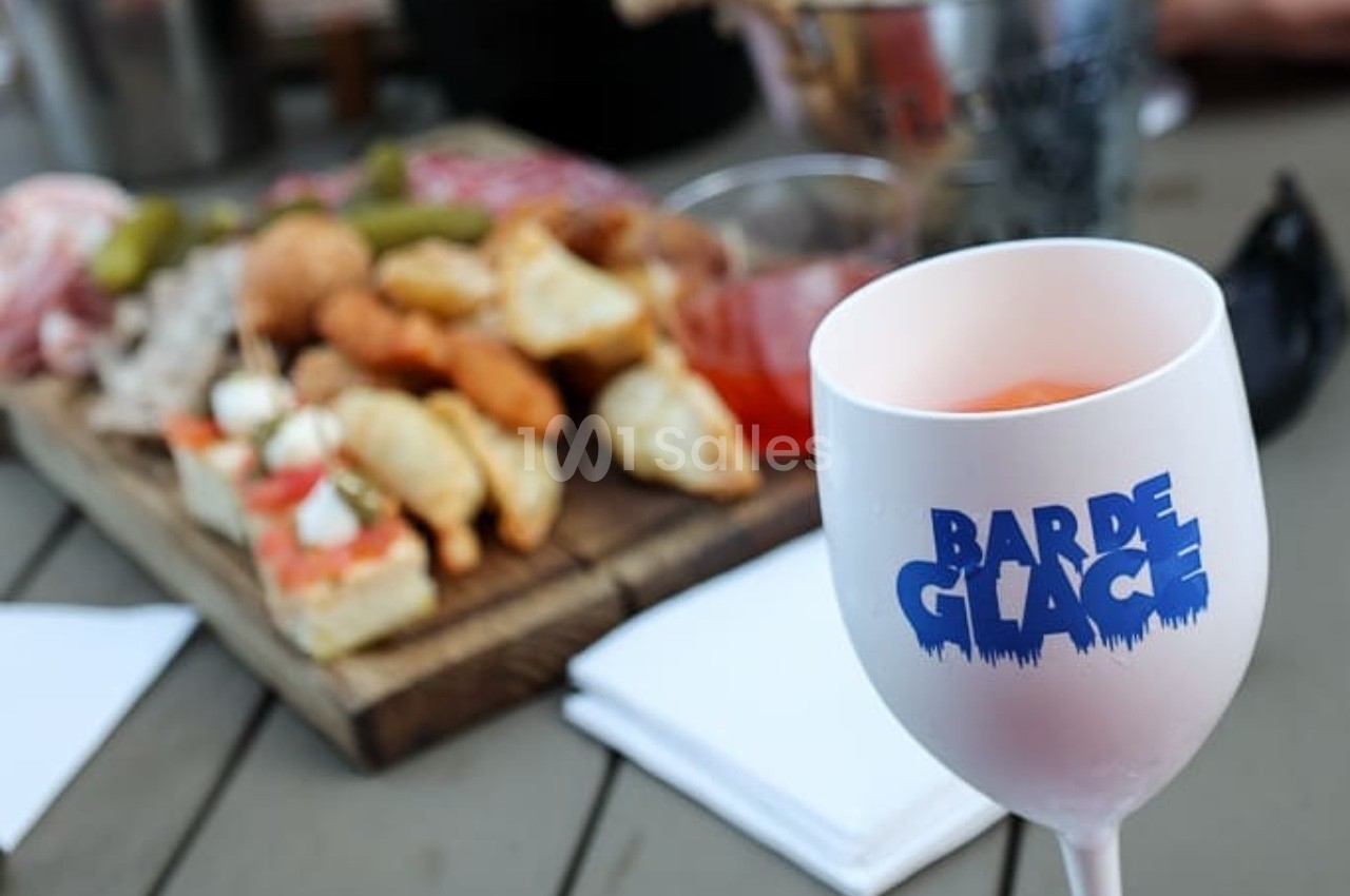 Verre de boisson marqué ’Bar de Glace’ au premier plan, plateau de bouchées variées flou en arrière-plan.