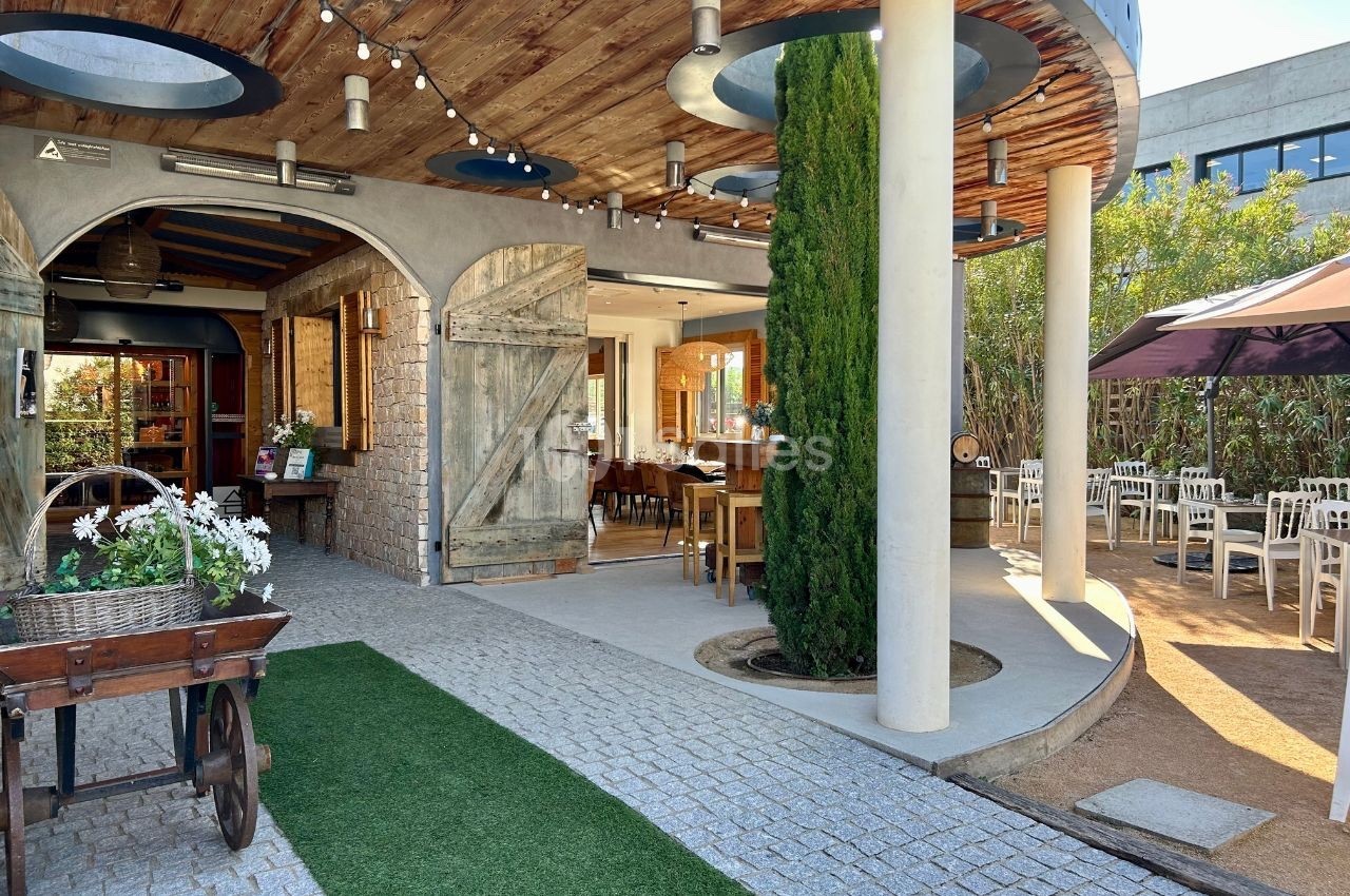 Entrée d'un restaurant avec terrasse extérieure, mobilier en bois, plantes et éclairage suspendu sous un toit en bois.