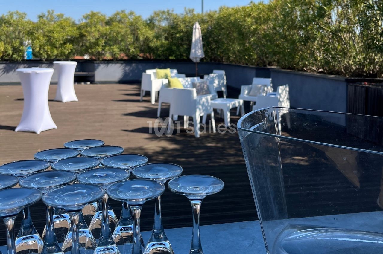 Terrasse ensoleillée avec tables hautes, chaises blanches et verres empilés au premier plan.