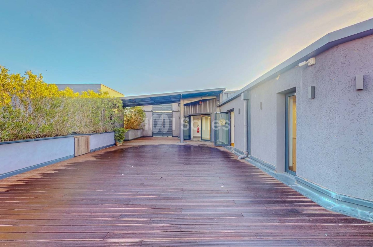 Terrasse en bois avec végétation sur le côté, entourée de bâtiments modernes sous un ciel dégagé.
