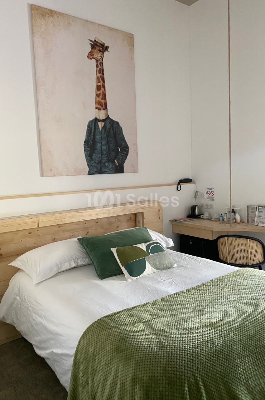 Chambre lumineuse avec lit double, tête de lit en bois, plaid vert, tableau mural et coin bureau avec chaise en rotin.