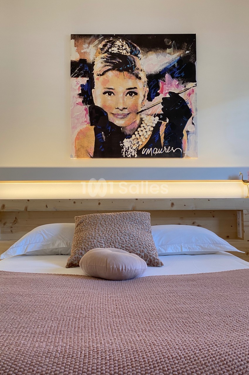 Lit double avec couverture rose, coussins assortis et tableau coloré représentant un visage féminin au mur.