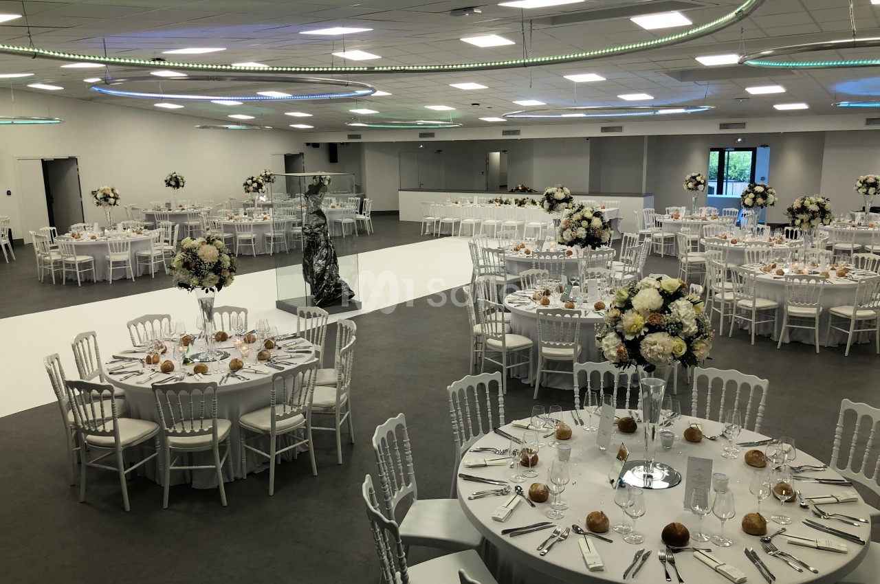 Salle de réception élégante avec tables rondes dressées, décorations florales et éclairage moderne.
