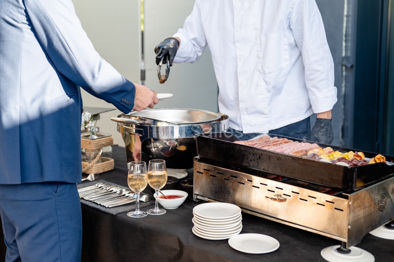 Un chef sert des plats chauds à un buffet avec des grillades, des assiettes et des verres de vin sur la table.
