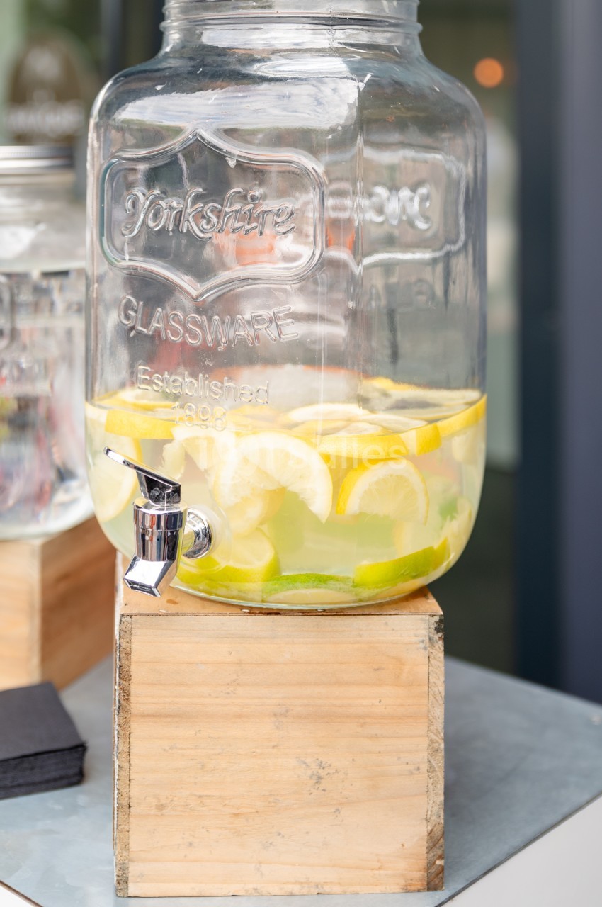 Distributeur en verre rempli d'eau infusée avec des tranches de citron et de citron vert, posé sur un support en bois.