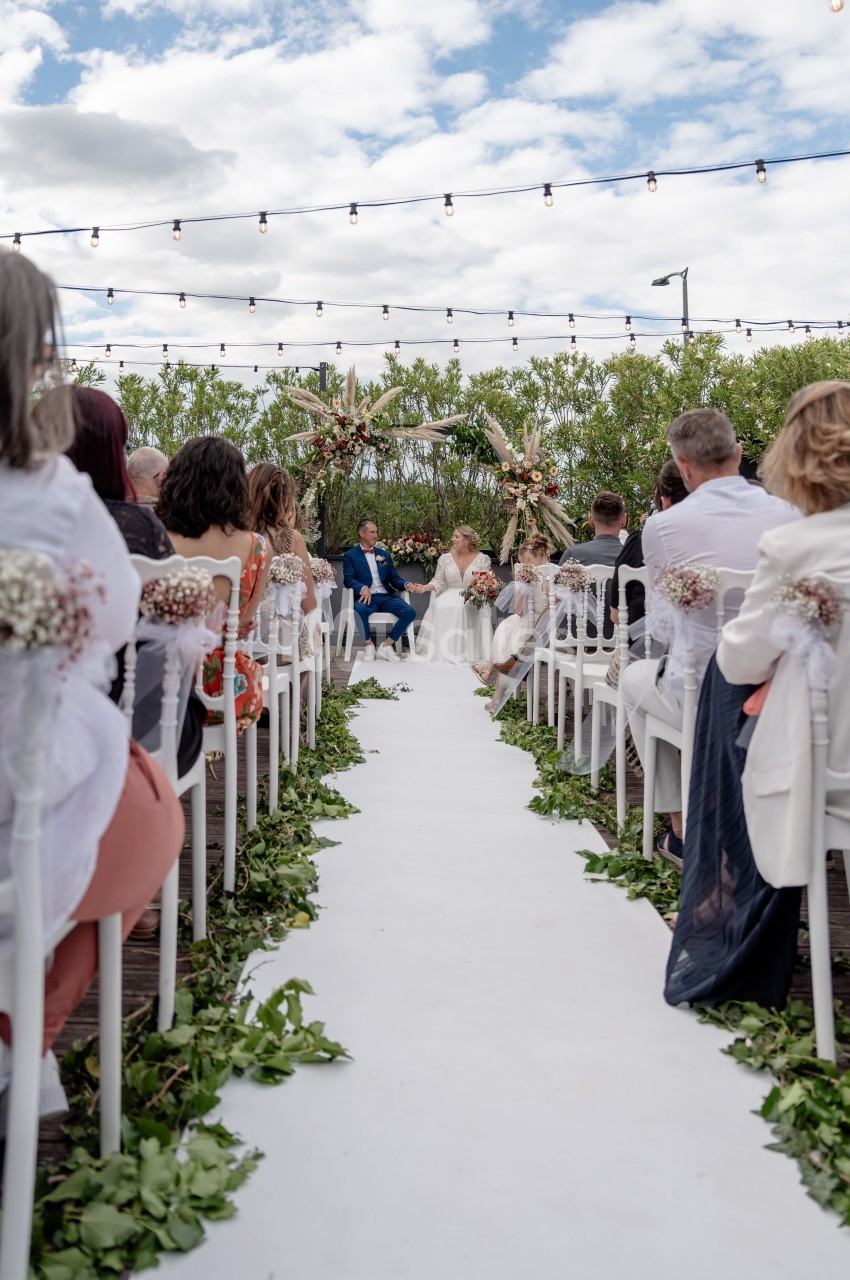 Allée centrale décorée de feuillages menant à une cérémonie de mariage en extérieur sous un ciel partiellement nuageux.
