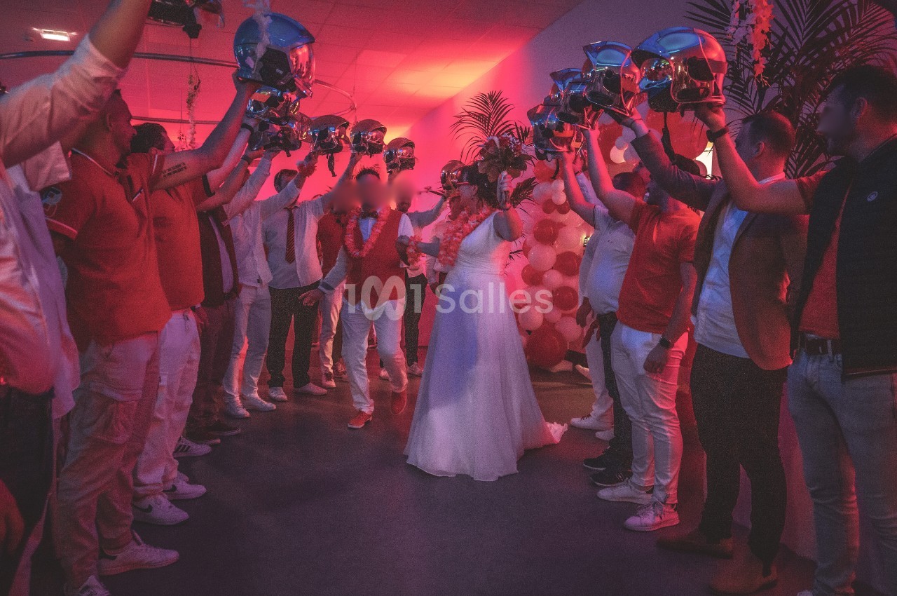 Un couple marche sous une haie d'honneur formée par des personnes levant des casques dans une salle éclairée en rouge.