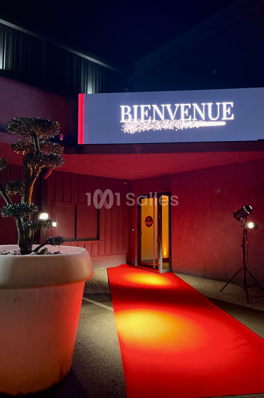 Entrée éclairée avec tapis rouge, panneau ’Bienvenue’ lumineux et plante décorative dans un pot blanc.