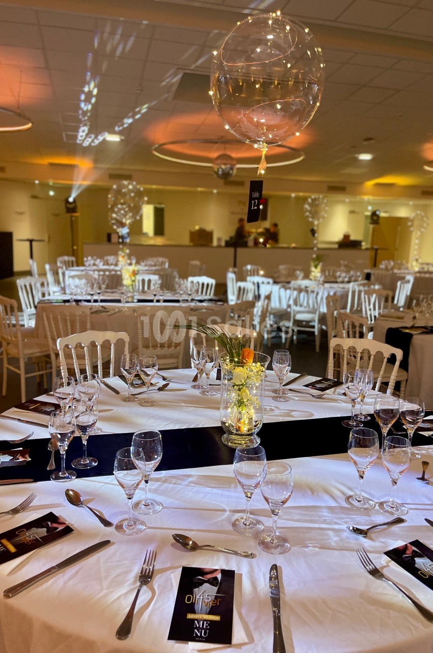 Salle de réception décorée avec des tables rondes dressées, centre de table floral et ballons suspendus.