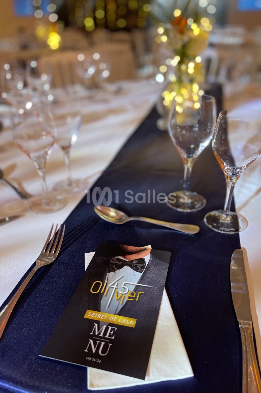 Table élégamment dressée avec nappe bleue, verres, couverts et un menu posé sur une serviette blanche.