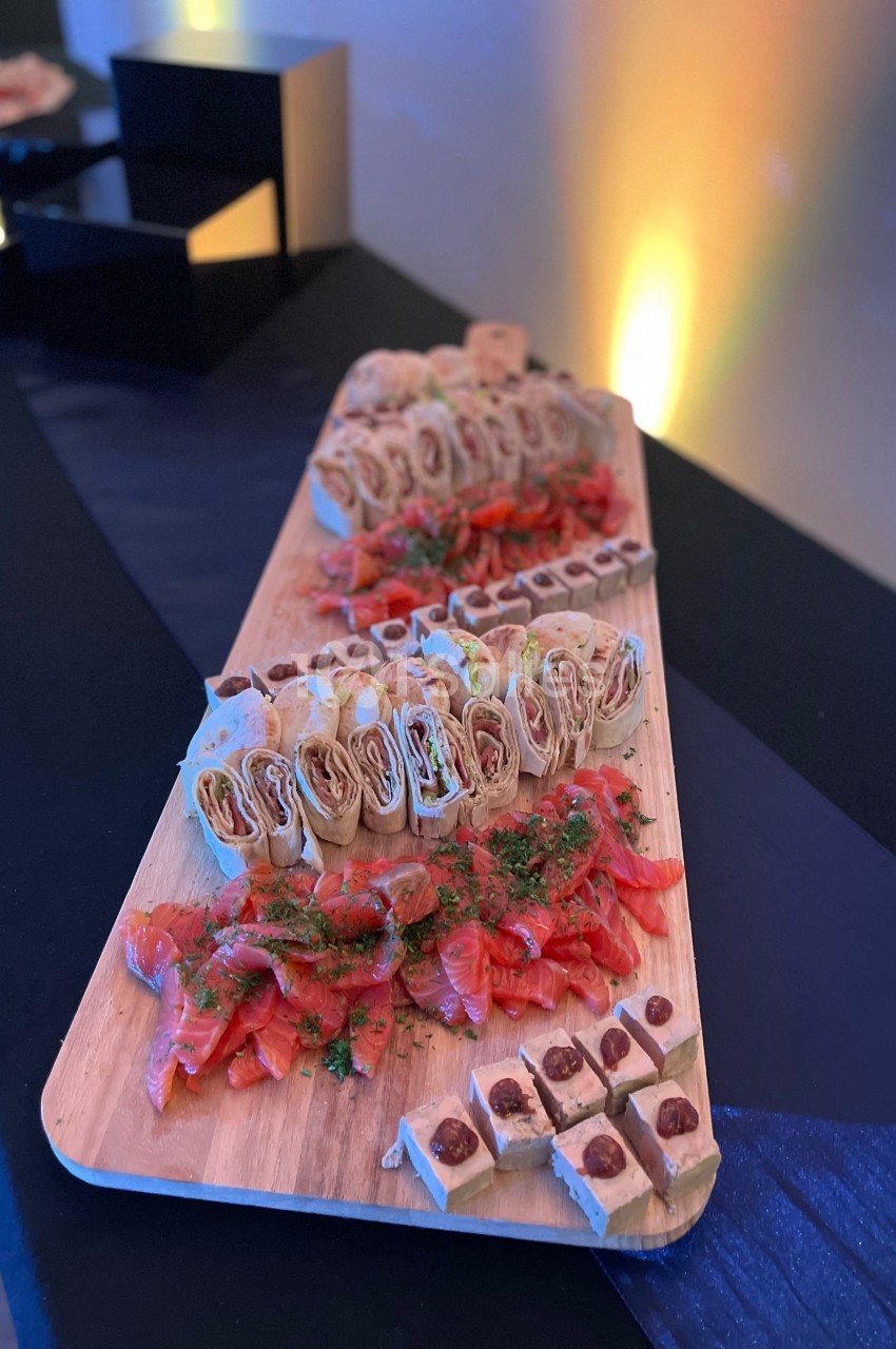 Plateau en bois avec des roulés apéritifs, du saumon fumé et des bouchées sucrées, présenté sur une table noire.