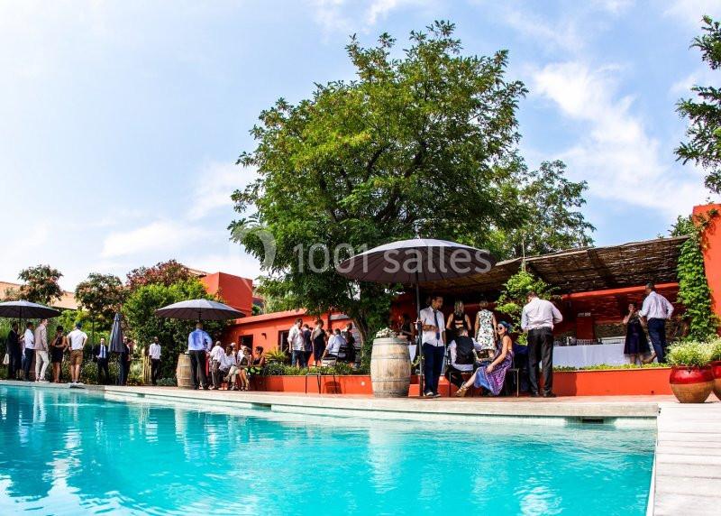 Personnes rassemblées près d'une piscine dans un cadre extérieur avec arbres, parasols et bâtiments colorés.