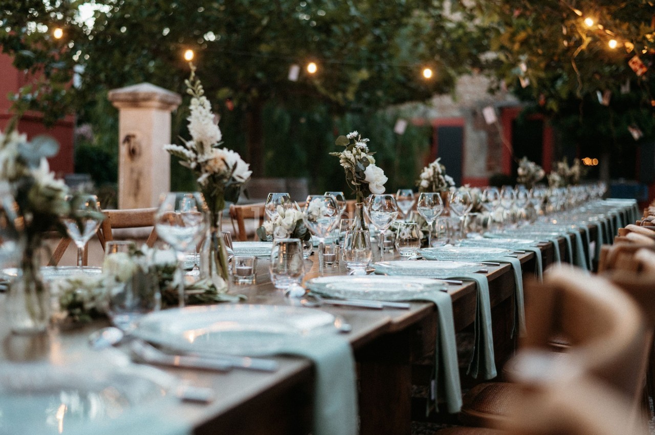 Table dressée en extérieur avec nappes bleues, verres, couverts et décorations florales sous des guirlandes lumineuses.