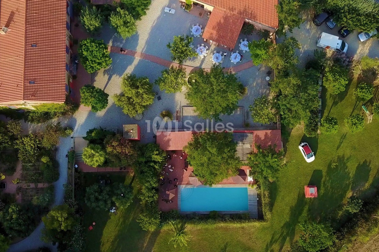 Vue aérienne d'une propriété avec piscine, terrasses, arbres et bâtiments aux toits rouges dans un cadre verdoyant.