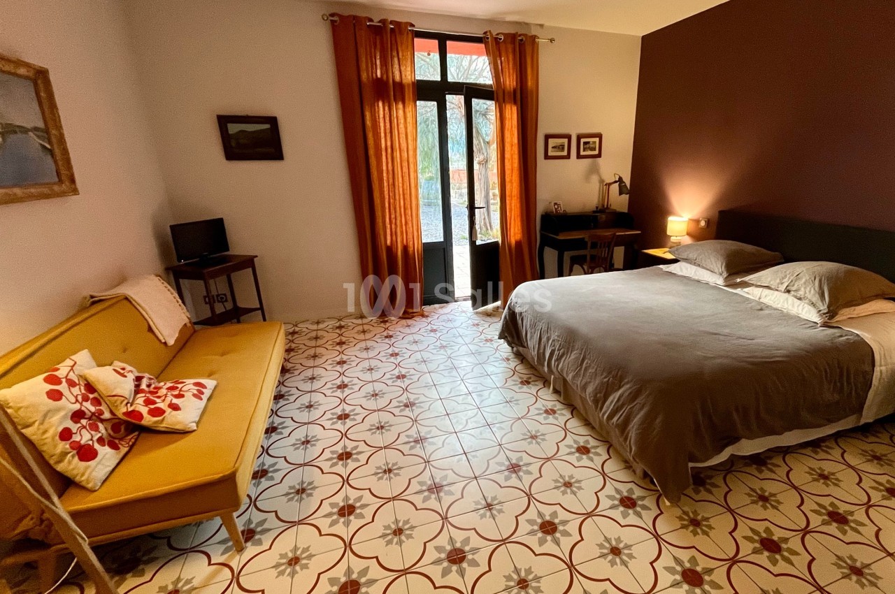 Chambre lumineuse avec lit double, canapé jaune, bureau, sol en carreaux décoratifs et porte-fenêtre donnant sur l'extérieur.