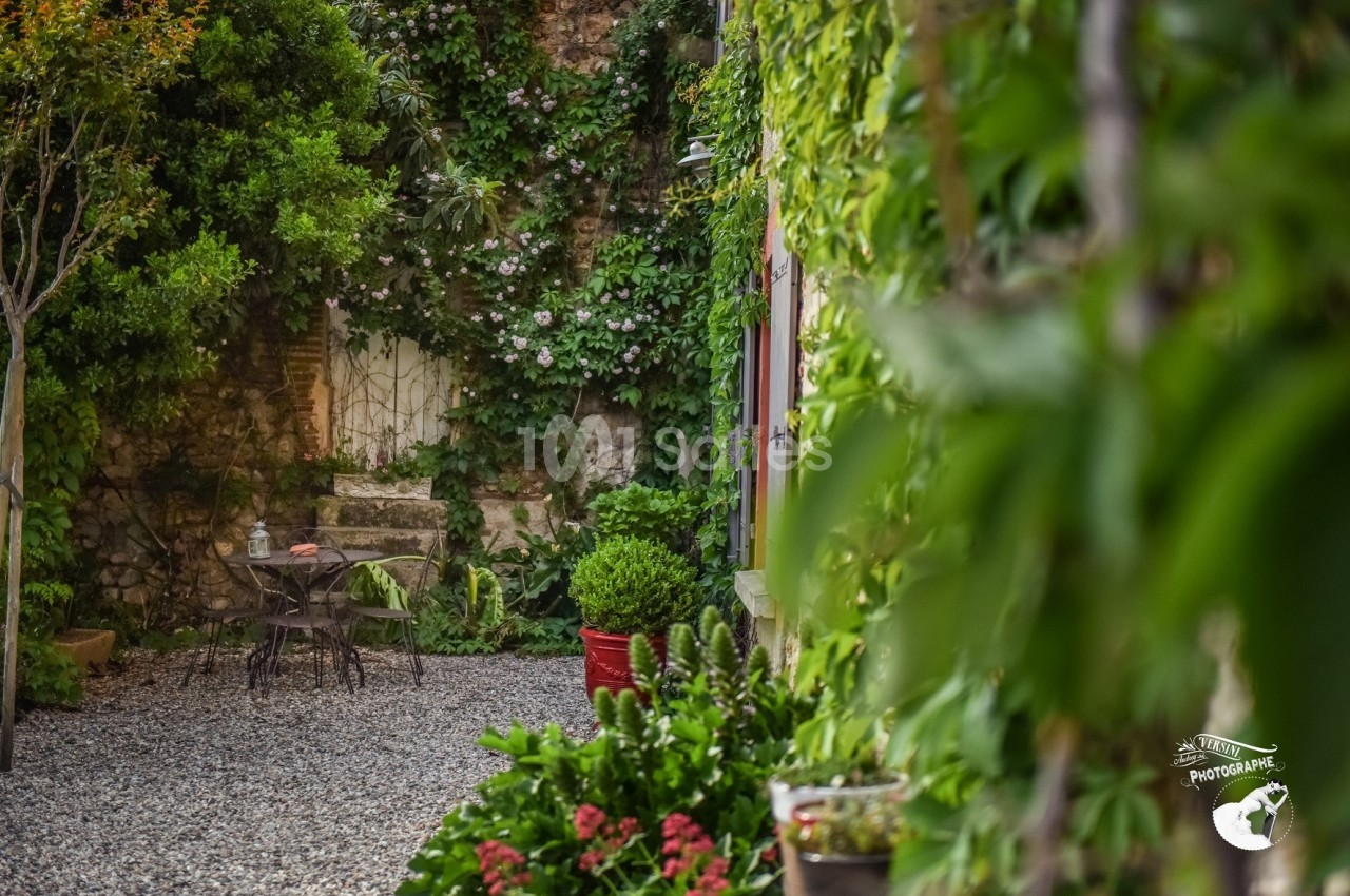Cour intérieure avec table et chaises en métal, entourée de plantes grimpantes et de pots de fleurs sur un sol en gravier.