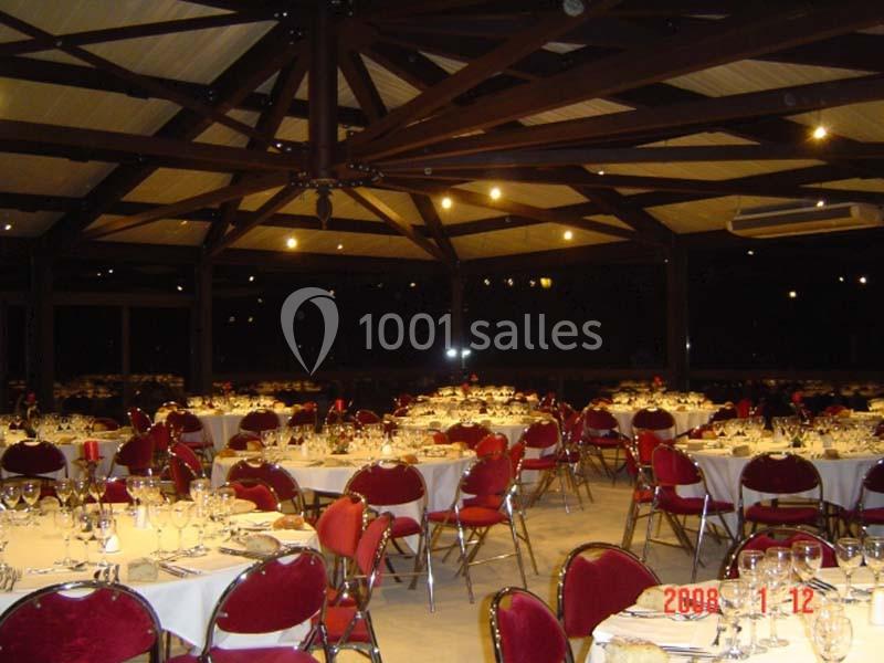 Salle de réception avec tables rondes dressées, nappes blanches, chaises rouges et éclairage chaleureux.