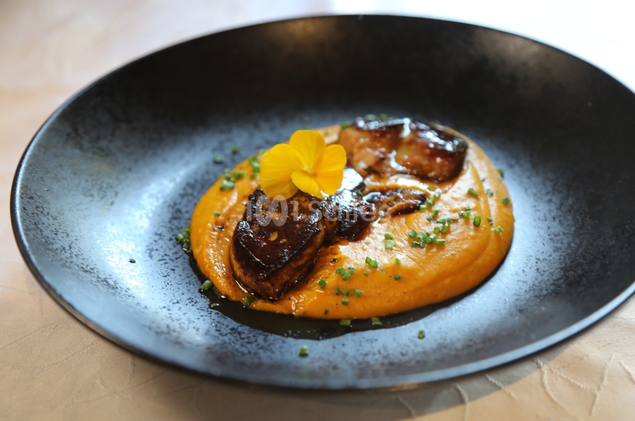 Assiette de purée de légumes orange garnie de morceaux de foie gras poêlé, ciboulette et une fleur comestible.