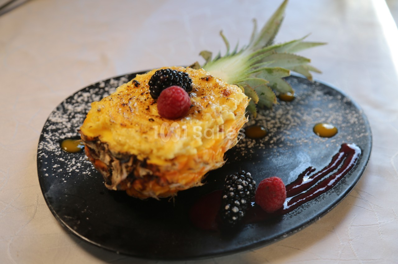 Demi-ananas garni d'un gratin sucré, décoré de fruits rouges et de coulis, présenté sur une assiette noire.