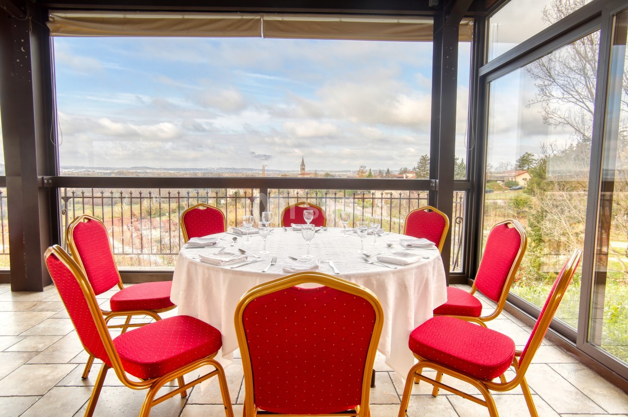 Salle lumineuse avec une table ronde dressée, entourée de chaises rouges, offrant une vue panoramique sur l'extérieur.