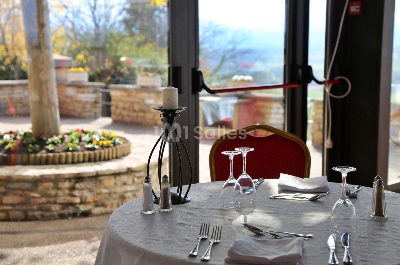 Table dressée avec nappe blanche, verres, couverts et bougeoir, près d'une baie vitrée donnant sur une terrasse ensoleillée.