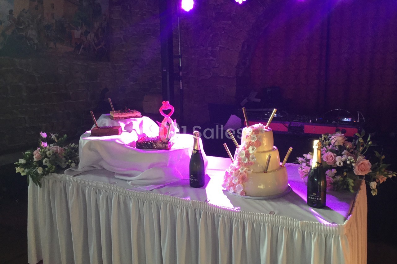 Table de fête décorée avec des fleurs, des bouteilles de champagne et des gâteaux, éclairée par une lumière rose.