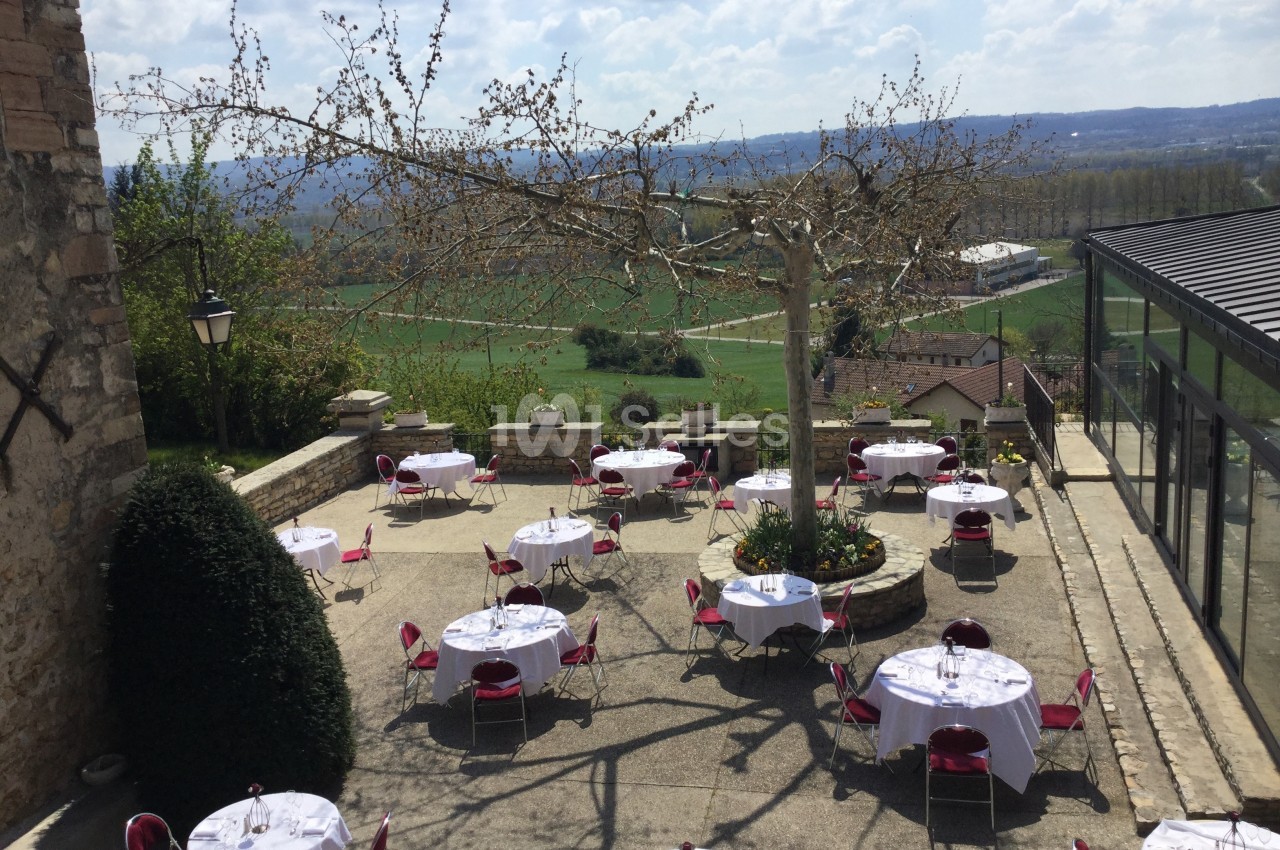 Terrasse extérieure avec tables dressées, chaises rouges, arbre central et vue sur une campagne vallonnée.