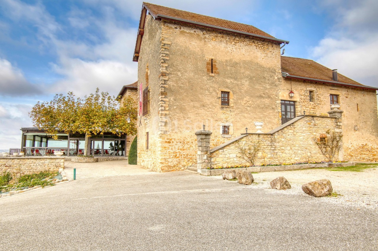 Bâtiment en pierre ancienne avec toit en tuiles, entouré d'arbres et d'une terrasse moderne sous un ciel partiellement…