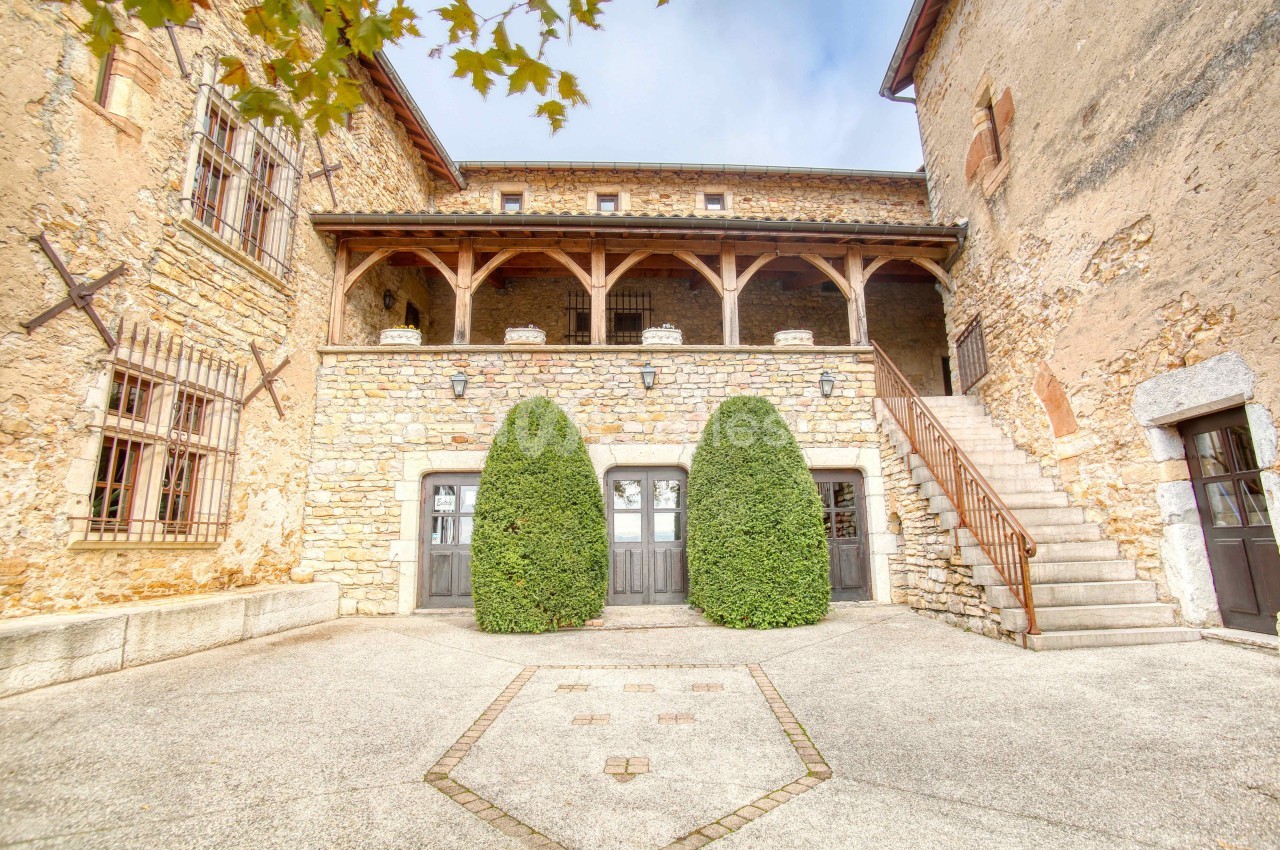 Façade en pierre d'un bâtiment ancien avec balcon en bois, escaliers latéraux et deux arbustes taillés devant.