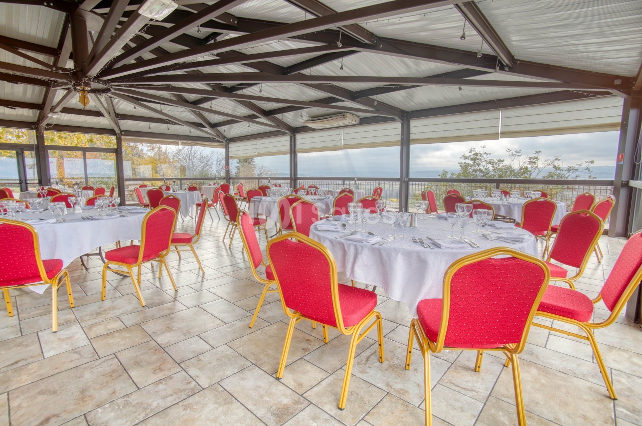 Salle lumineuse avec tables rondes dressées de nappes blanches et chaises rouges, entourée de baies vitrées.
