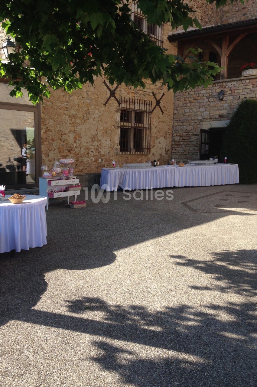 Cour extérieure d'un bâtiment en pierre avec des tables décorées pour un événement, partiellement ombragée par des arbres.