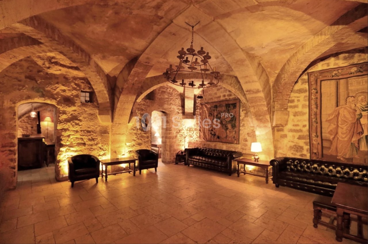 Salle voûtée en pierre éclairée par des lampes, avec canapés en cuir, tapisseries murales et mobilier en bois sombre.