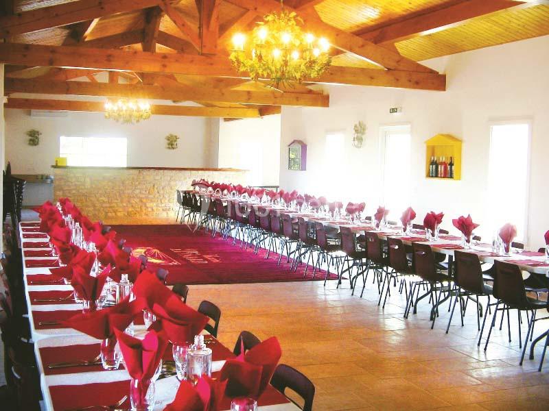 Salle de réception lumineuse avec deux longues tables dressées, nappes blanches et serviettes rouges, sous un plafond en…