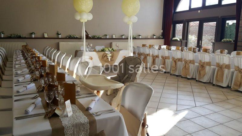 Salle de réception décorée avec des tables alignées, nappes blanches, rubans beige et ballons suspendus.