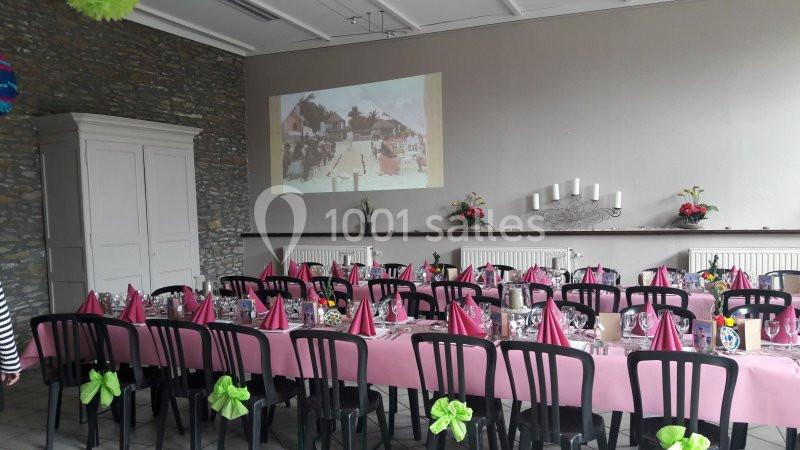 Salle décorée pour un repas festif avec tables dressées, nappes roses, serviettes pliées et projection murale en arrière…