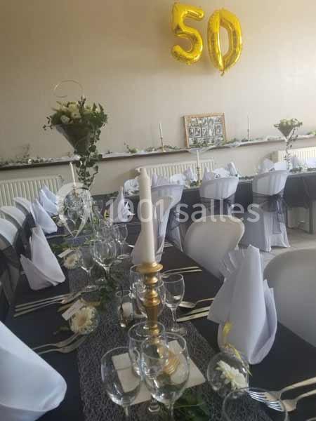 Salle décorée pour une fête avec tables dressées, nappes noires et blanches, bougies et ballons dorés ’50’ accrochés au mur.
