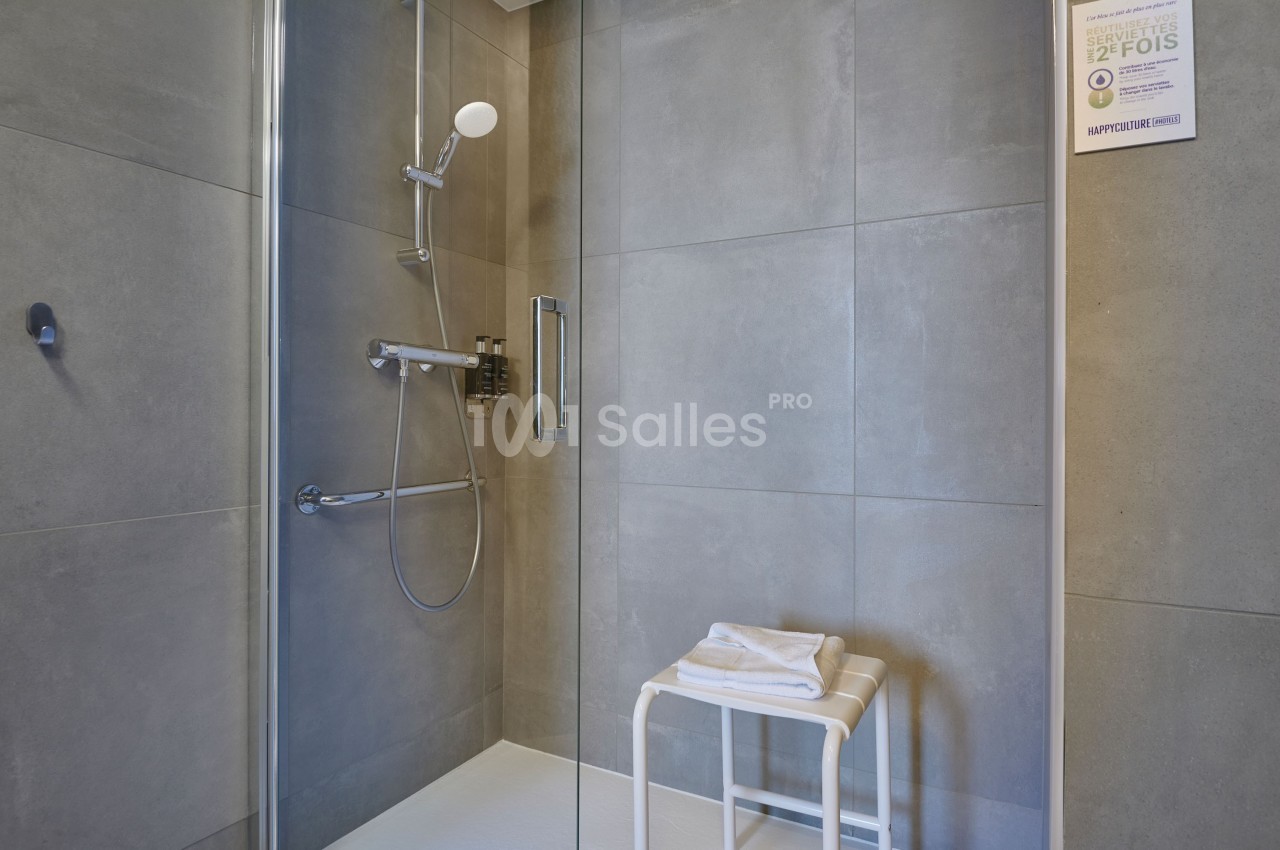 Douche moderne avec parois en verre, carrelage gris, pommeau mural et tabouret blanc posé au sol.