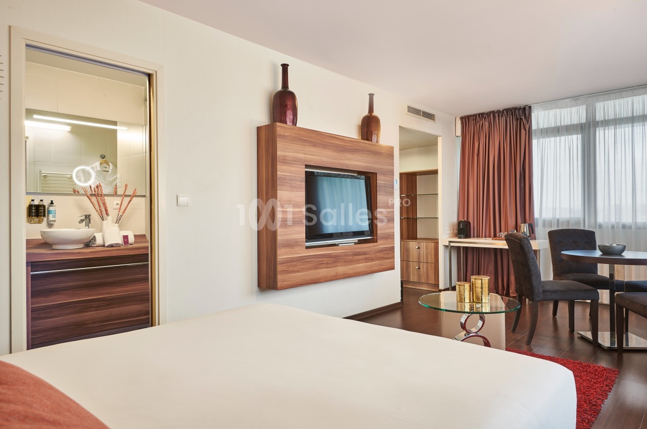 Chambre moderne avec lit double, télévision murale, coin repas, et salle de bain visible à travers une porte ouverte.