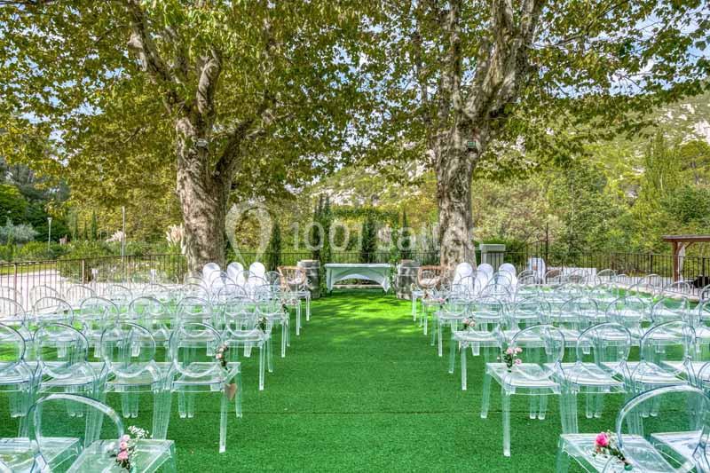 Chaises transparentes disposées en rangées sur une pelouse verte, encadrées par deux grands arbres, pour une cérémonie en…