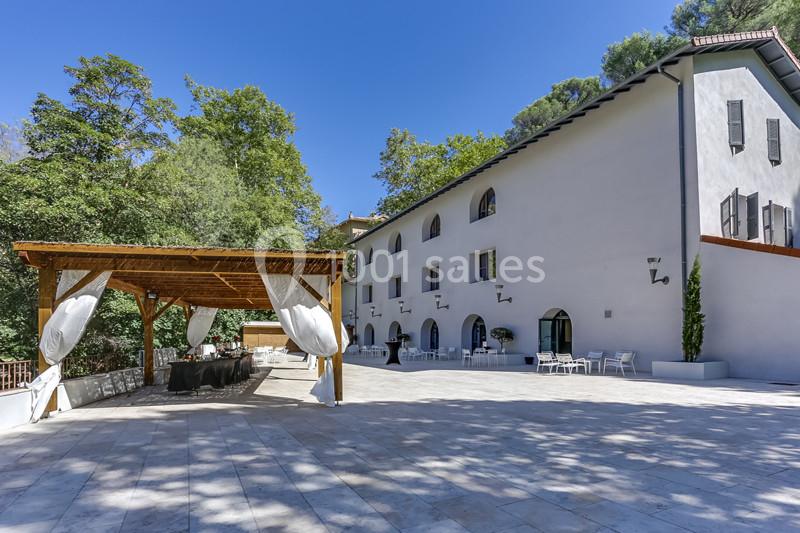 Grande terrasse pavée avec pergola en bois, tables et chaises blanches, devant un bâtiment blanc entouré de végétation.