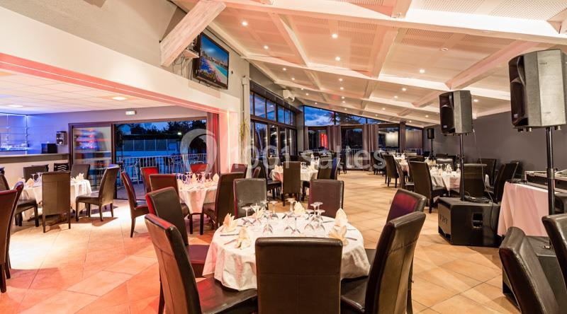 Salle de restaurant lumineuse avec tables rondes dressées, chaises en cuir foncé et grandes baies vitrées donnant sur l…