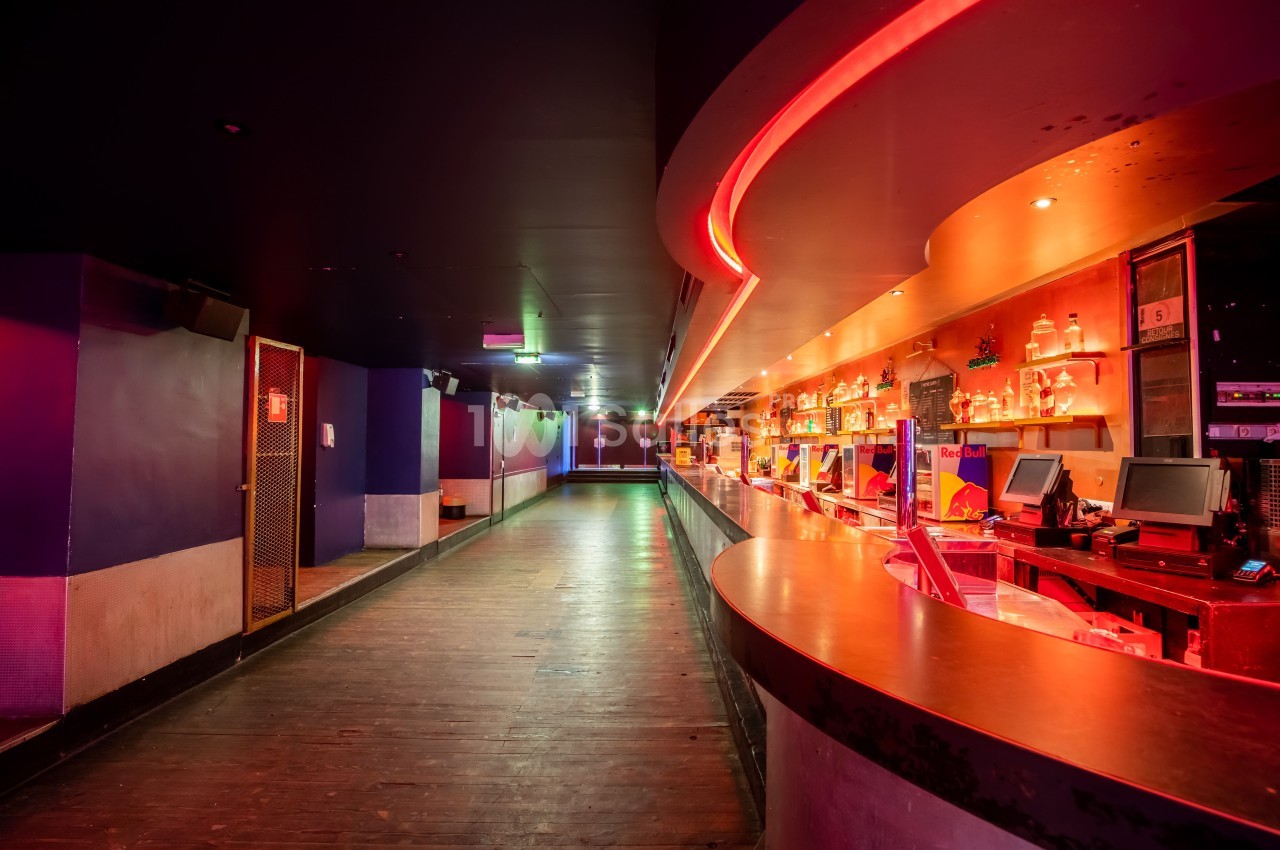 Intérieur d'une discothèque avec un long bar éclairé de néons rouges et une piste de danse vide en arrière-plan.