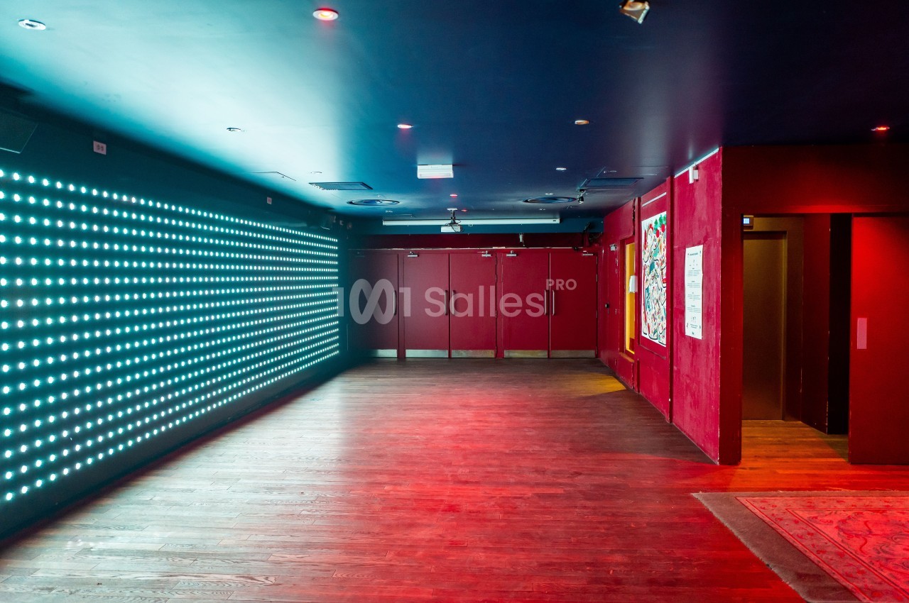 Couloir avec murs rouges, sol en bois, panneau lumineux à gauche et portes vitrées au fond.