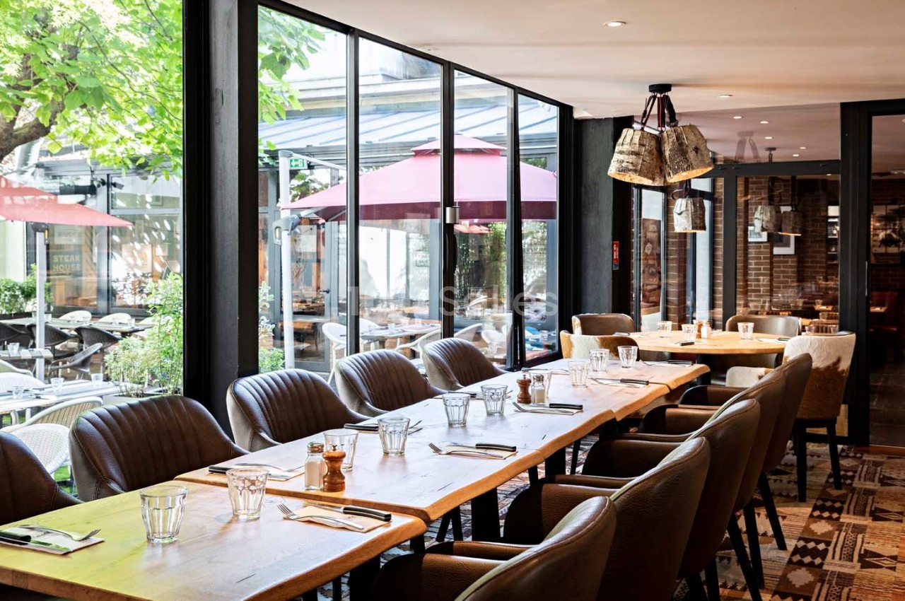 Salle de restaurant lumineuse avec de grandes baies vitrées donnant sur une terrasse équipée de parasols rouges.