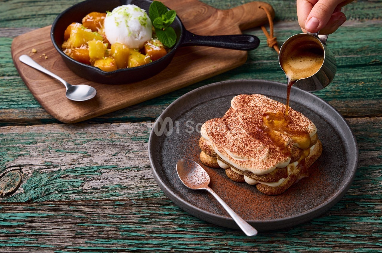 Une main verse du café sur un tiramisu, avec en arrière-plan un dessert aux fruits et glace dans une poêle.