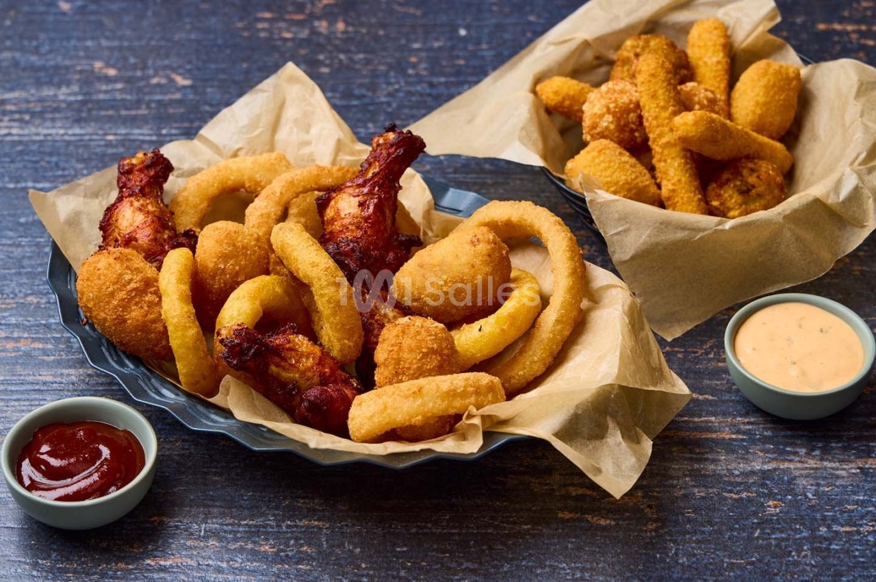 Assortiment de snacks frits comprenant ailes de poulet, rondelles d'oignon et sauces, présenté sur du papier sulfurisé.