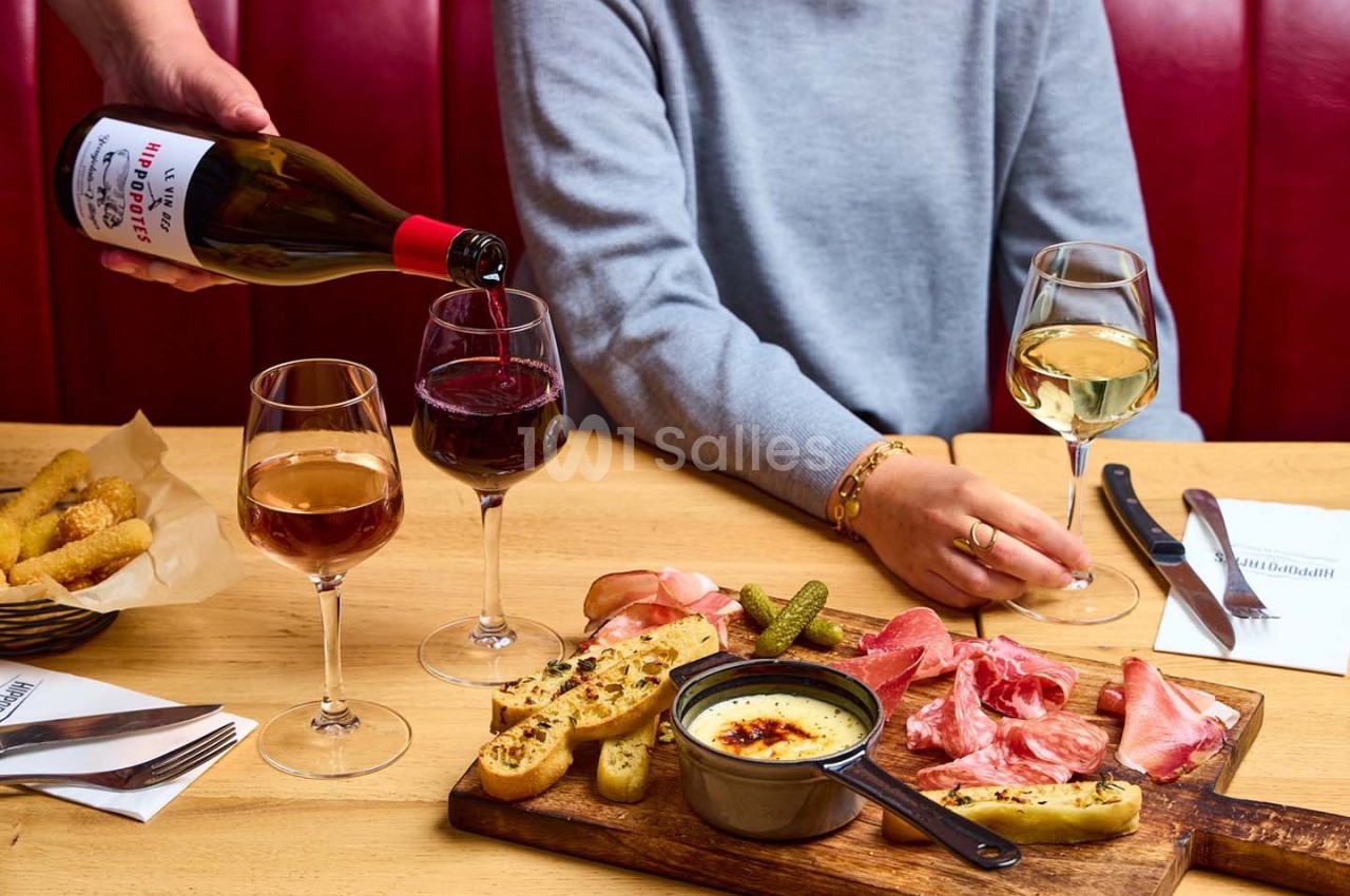 Un serveur verse du vin rouge dans un verre sur une table garnie de charcuterie, fromage fondu et autres accompagnements.