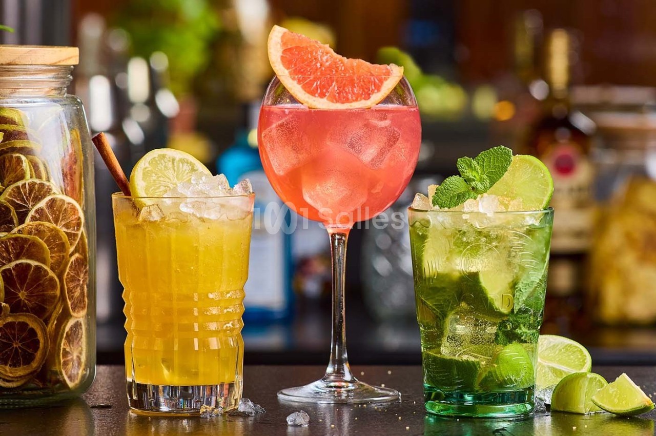 Trois cocktails colorés garnis de fruits frais, disposés sur un bar avec des ingrédients en arrière-plan.