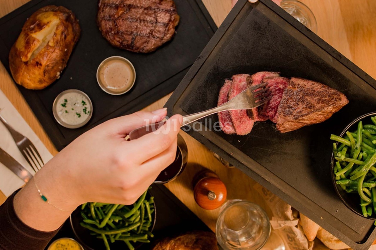 Une personne coupe un morceau de viande saignante sur une planche, accompagnée de haricots verts et d'autres plats.