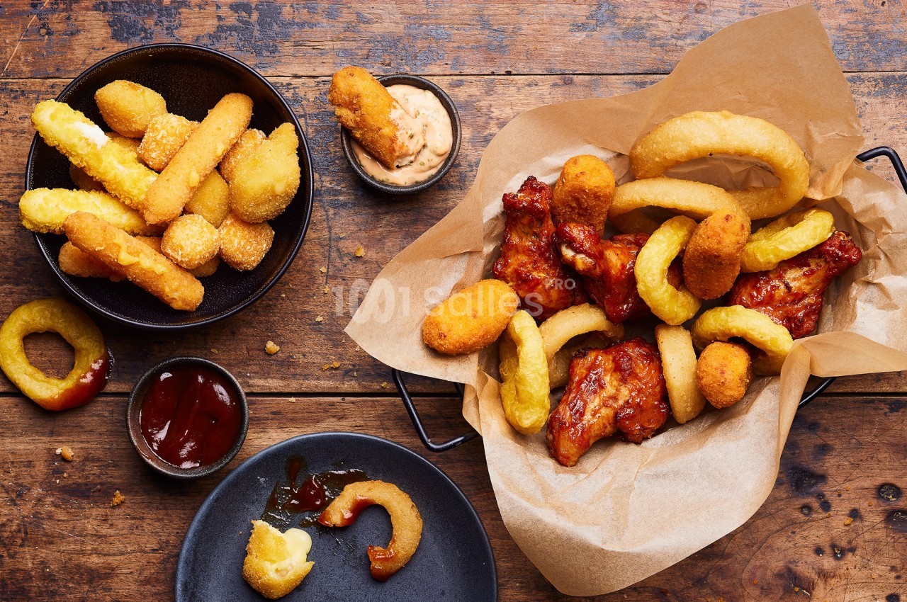 Assortiment de bouchées frites, ailes de poulet, rondelles d'oignon et sauces sur une table en bois.